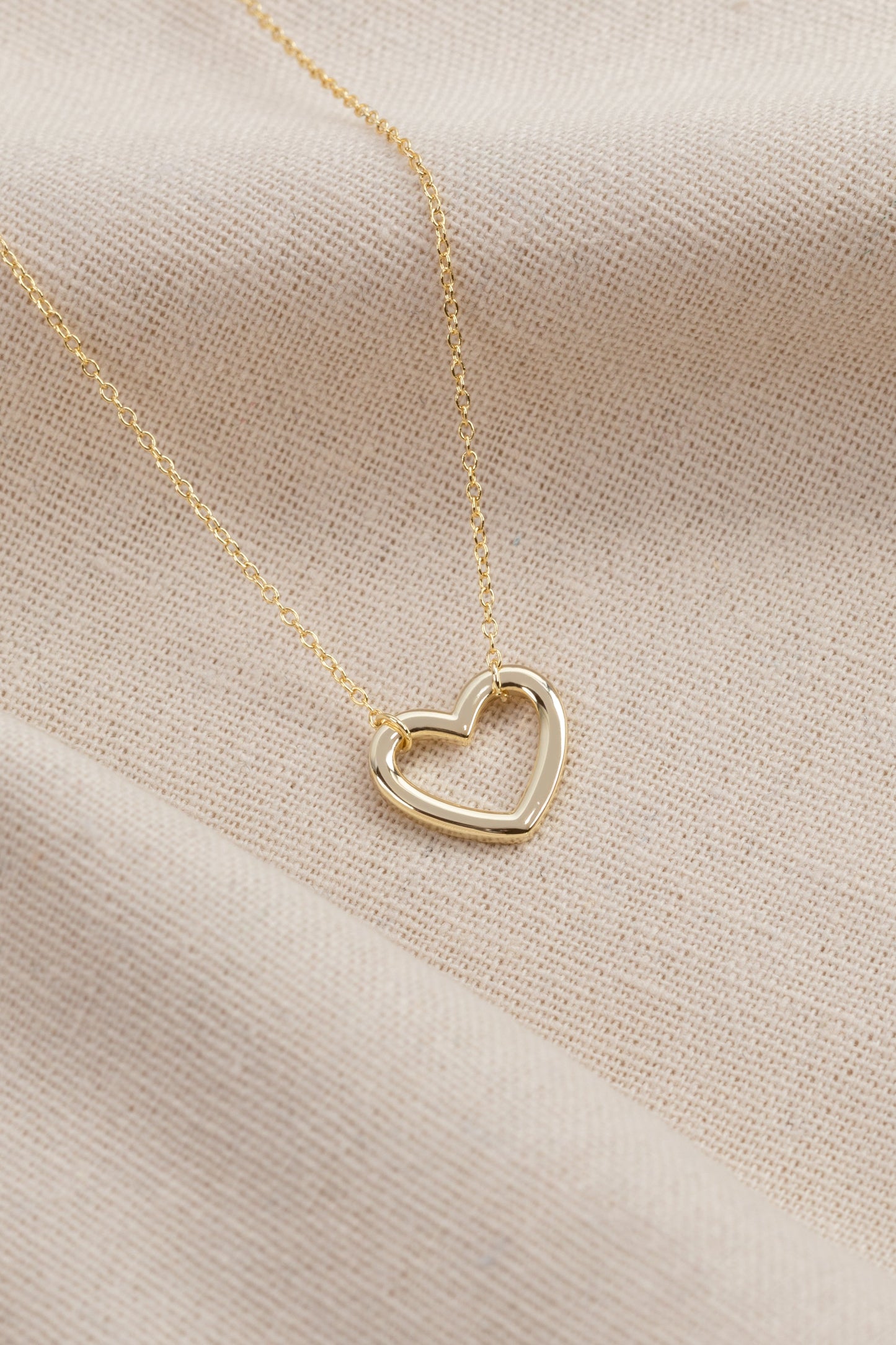 gold plated heart shaped pendant on a gold plated chain . this necklace is on a hessian style material backdrop