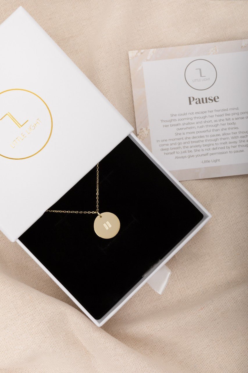 Gold pendant on a gold necklace with a pause symbol on disc. its displayed in a white box with a black foam insert
